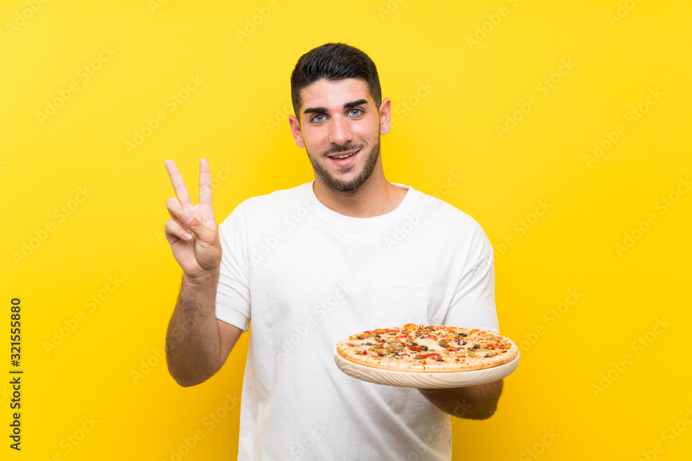 Young handsome man holding a pizza over isolated yellow wall smiling and showing victory sign Young handsome man holding a pizza over isolated yellow wall smiling and showing victory sign