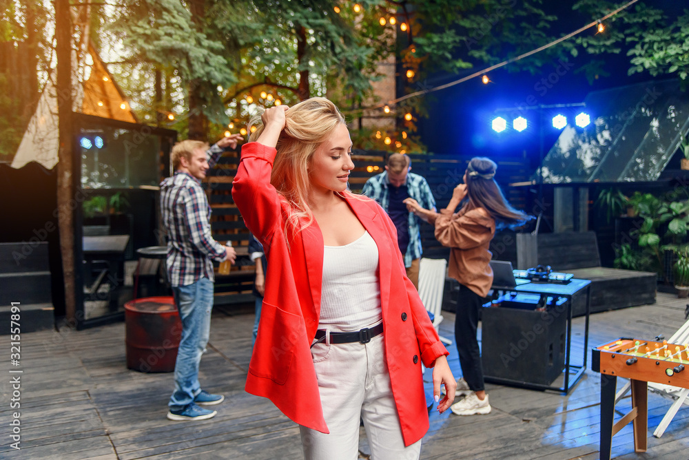 Close up of attractive blond girl with blue eyes posing to the camera with her friends dancing in disco light on the background. Outdoors evening party concept. Close up of attractive blond girl with blue eyes posing to the camera with her friends dancing in disco light on the background. Outdoors evening party concept.