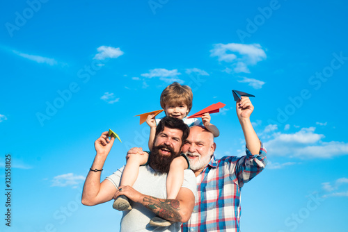 Kid pilot with toy jetpack against sky background. Happy three generations of men have fun and smiling on blue sky background. Happy child playing outdoors. © Volodymyr
