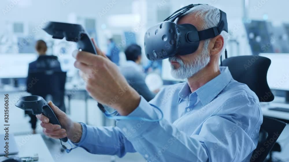 Industrial Design Engineer Wearing Virtual Reality Headset and Holding Controllers, Uses VR technology for Industrial Design, Development using Computer. Modern Factory Office, Workshop with Machinery Industrial Design Engineer Wearing Virtual Reality Headset and Holding Controllers, Uses VR technology for Industrial Design, Development using Computer. Modern Factory Office, Workshop with Machinery