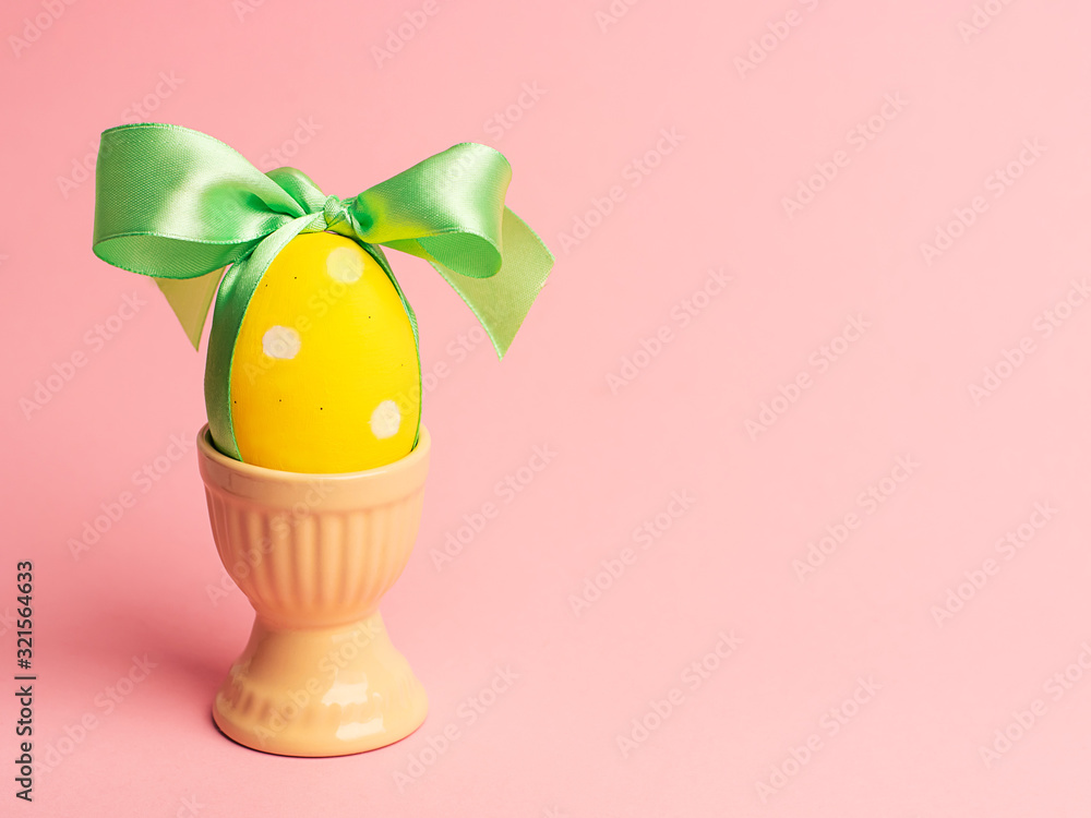 Easter creative concept. One yellow egg tied with a green ribbon standinng in the yellow egg cup a on a pink background. Minimalism, copy space, close up