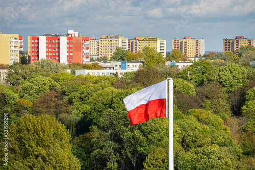 Polish state flag in the background of sunlit green trees in autumn. In the distance is a row of colorful new high-rise houses and a blue sky with clouds. View in Lublin, Poland