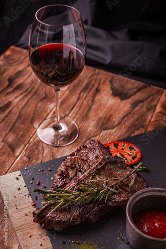 Cooked meat t-bone steak on serving board with garlic cloves, tomatoes, rosemary, spices and glass of red wine over rustic wooden background