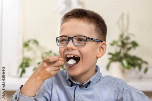 Uśmiechnięty chłopiec w wieku szkolnym siedzi przy stole i wkłada do ust łyżeczkę z białym cukrem.