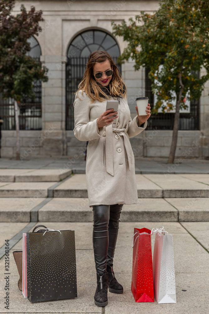 Fototapeta premium Attractive slim woman looks at her mobile phone while having a coffee in the street next to her shopping bags