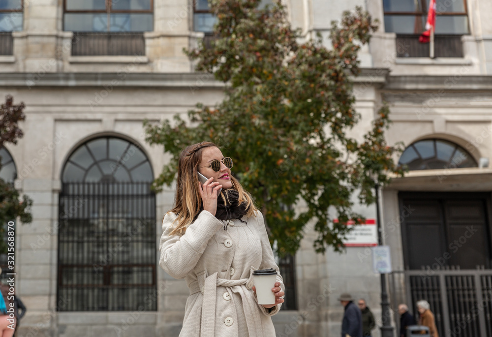 Attractive blonde business woman with white coat and black glasses speaks with her smartphone while having a coffee