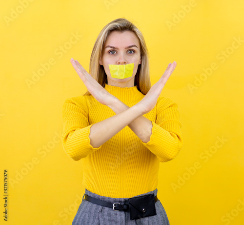 Portrait of a blonde girl with crossed hands gesture and yellow tape taped her mouth. Censorship concept on yellow background. Stop, warning
