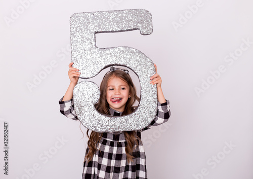 Birthday party. Grimacing little baby shows tongue. Happy child with number 5. Smiling little girl at studio background. Girl  holding in his hands figure of five. Sale.