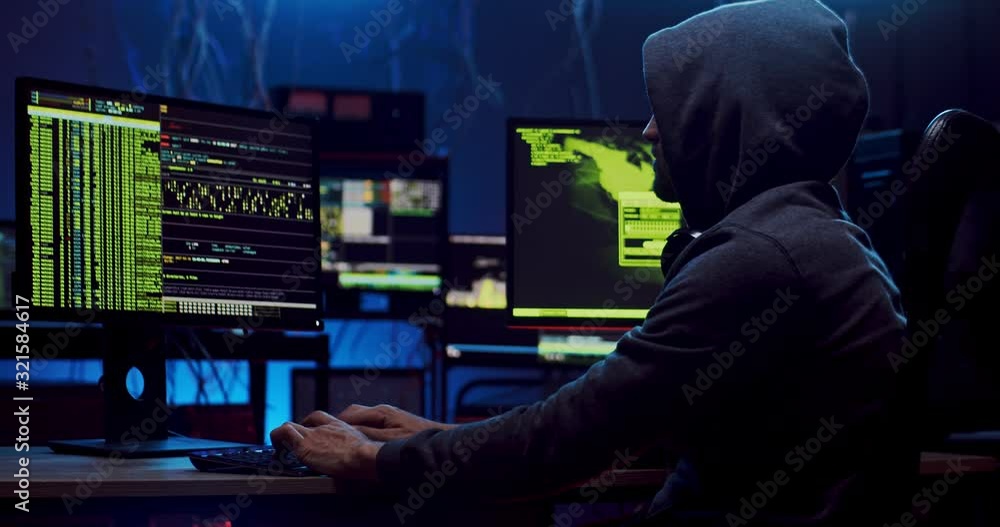 Side view on the male anonymous hacker in the hood creating virus while coding at the computer in the dark software room with digits running on the screen. Side view on the male anonymous hacker in the hood creating virus while coding at the computer in the dark software room with digits running on the screen.