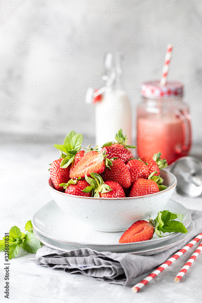 Fresh strawberries in a bowl and sprigs of mint on a white concrete table. Ingredient for smoothies. Bottle of milk, red glass mug and a blender. The recipe for the holiday Valentine's Day. Copy space Fresh strawberries in a bowl and sprigs of mint on a white concrete table. Ingredient for smoothies. Bottle of milk, red glass mug and a blender. The recipe for the holiday Valentine's Day. Copy space