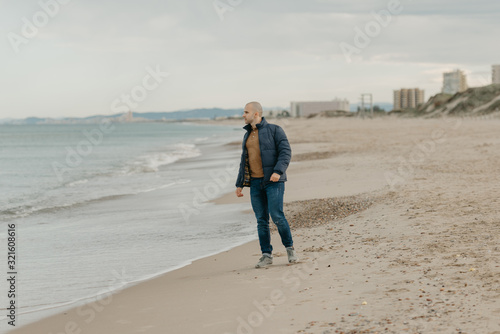 A muscular bald stylish man in the bologna jacket, jeans and sneakers poses on the shore of the Mediterranean Sea