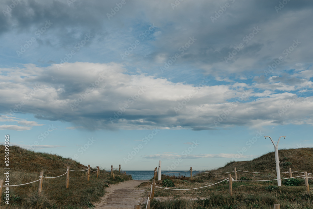 A walkway between sandy green dunes to the Mediterranean Sea. Clouds on the sky. A beach shower.