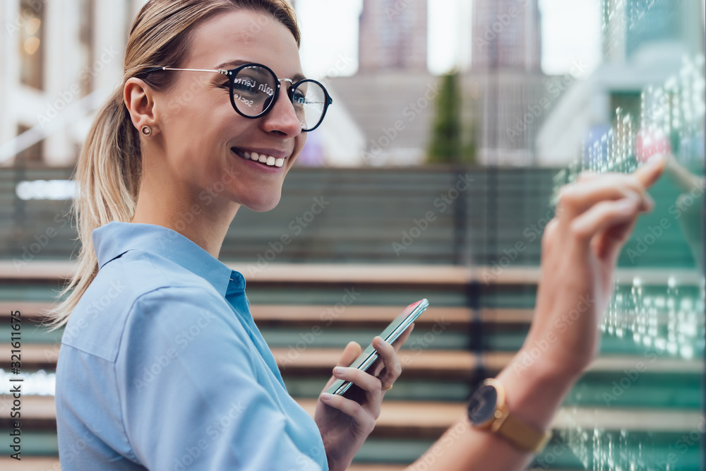 Fototapeta premium Interactive digital kiosk with public transport Info. Smiling female standing at big display with smartphone in hand.Young woman touching with finger interactive futuristic screen. Advanced technology