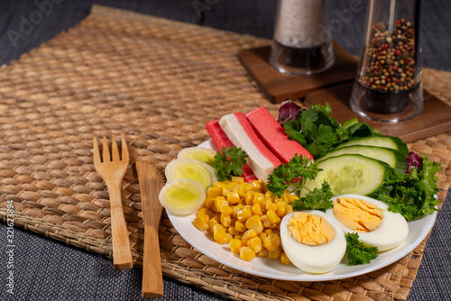 Salad plate on the table with wooden cutlery. Crab sticks, boiled egg, corn and greens. 