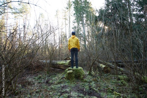 Man, weared in yellow raincoat, standing on a mossy stump among woods in forest