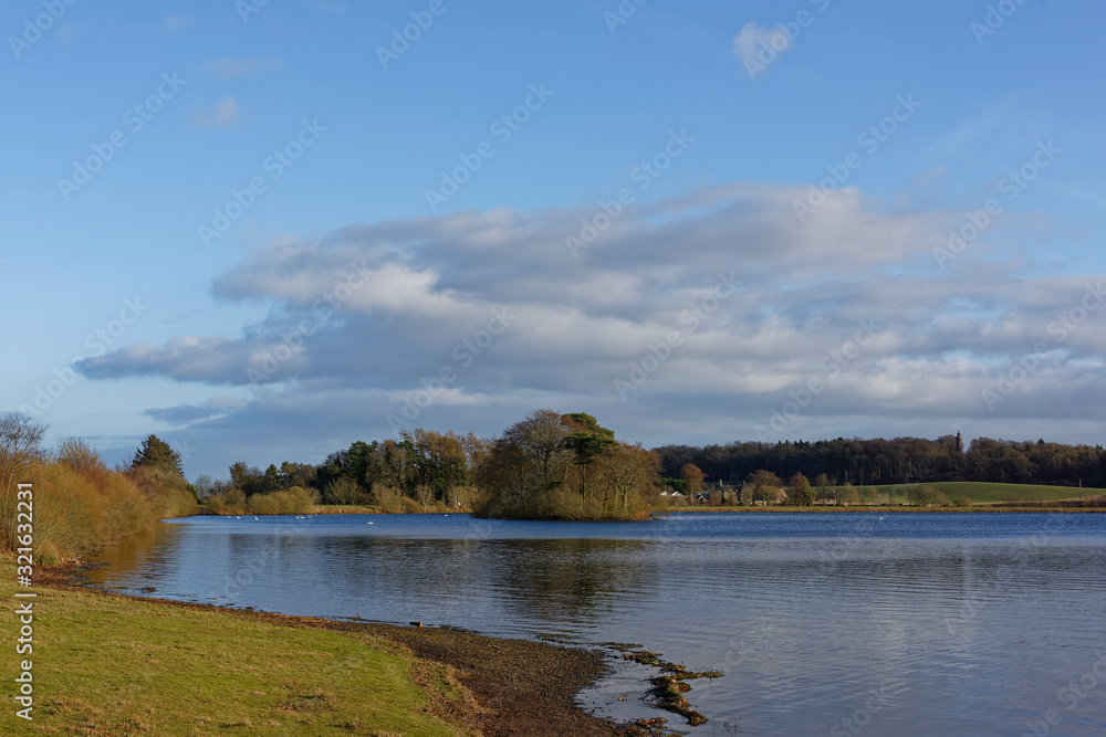 Looking over the main Reservoir at Monikie Country Park, with a heavily wooded small Island in the distance, and buildings and Fields in the background.