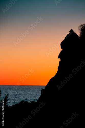 Silhouette of isolated cliff on sea coast with deep blue sea on orange sunset