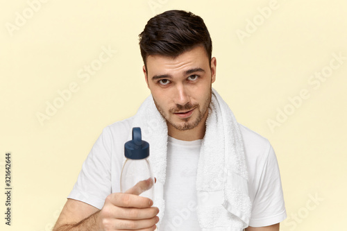 Isolated shot of handsome confident young European male with bristle posing with white towel around his neck, drinking water from plastic cup after physical training, going to take shower, smiling