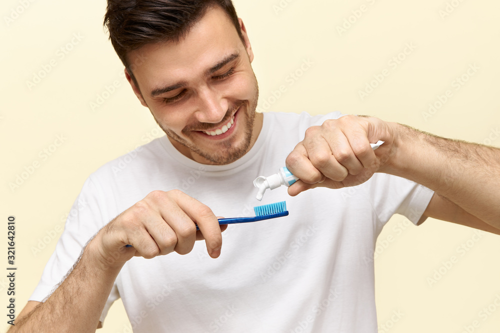 Close up of attractive young bearded man is brushing his teeth. He is standing and smiling. The man is squeezing tooth-paste into tooth-brush. Focus on cleaning tools Close up of attractive young bearded man is brushing his teeth. He is standing and smiling. The man is squeezing tooth-paste into tooth-brush. Focus on cleaning tools