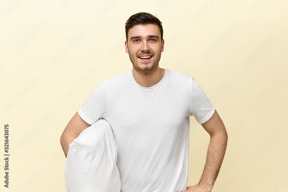 Bedding and bedtime concept. Isolated shot of attractive young male with bristle having tired look, smiling with relief being happy that he is going to sleep finally, holding pillow under his arm Bedding and bedtime concept. Isolated shot of attractive young male with bristle having tired look, smiling with relief being happy that he is going to sleep finally, holding pillow under his arm
