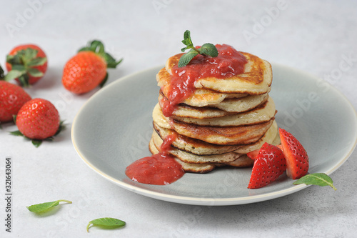 Pancakes with strawberry syrup. Fritters are decorated with fresh strawberries and mint leaves. Strawberries are scattered on the table surface. Light background.