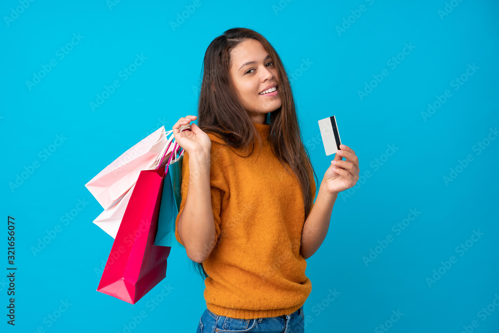Young Brazilian girl over isolated blue background holding shopping bags and a credit card Young Brazilian girl over isolated blue background holding shopping bags and a credit card