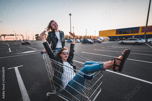 Two young happy women in sunglasses having fun shopping trolley race outdoors. Having fun at the mall parking lot at sunset