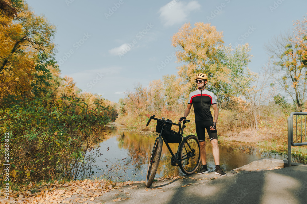 Obraz premium Portrait of man with bicycle standing on background of autumn landscape with river and looking to the side. Cyclist is resting on a bicycle by the lake. Cycling. concept