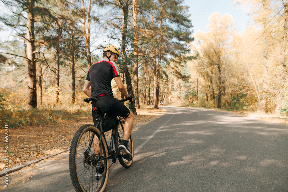 Back of a cyclist man in a sport bike suit and helmet stands on the road in the forest and looks away against the backdrop of an autumn landscape. Man rides a bicycle in a warm autumn day