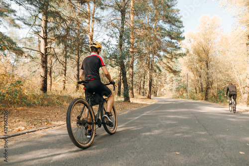 Cyclist's back stands on the road in the forest on a background of autumn landscape. A man in a helmet and cycling clothes sits on a bicycle and rests after a ride. Weekend bicycle hobby.