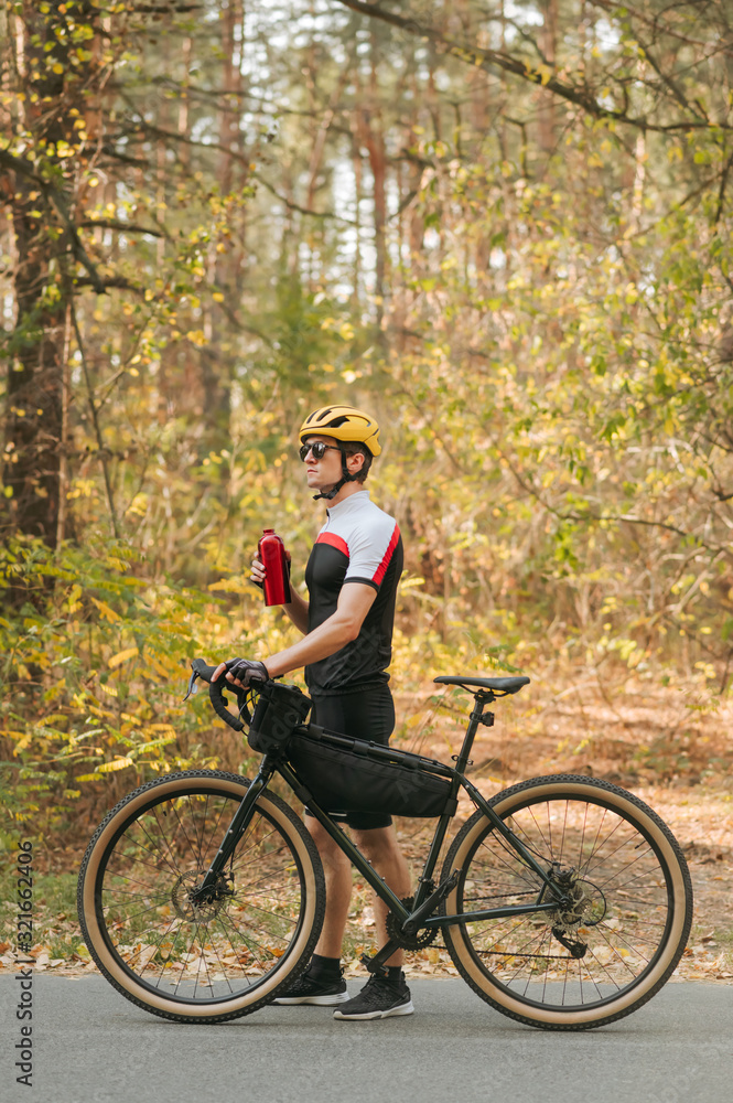 Portrait of cyclist walking with bicycle and red water bottle on road in forest and looking away on background of autumn trees. The cyclist rests after the ride, drinks water and stands by the bike