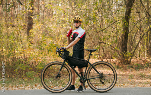 Athletic man stands on the road with a bicycle,background of autumn trees, looks in camera and drinks water from a red bottle.Thirsty bicyclist is resting, standing with a bicycle and bottle water © bodnarphoto