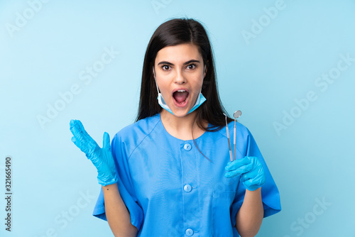 Young woman dentist holding tools over isolated blue background with surprise facial expression