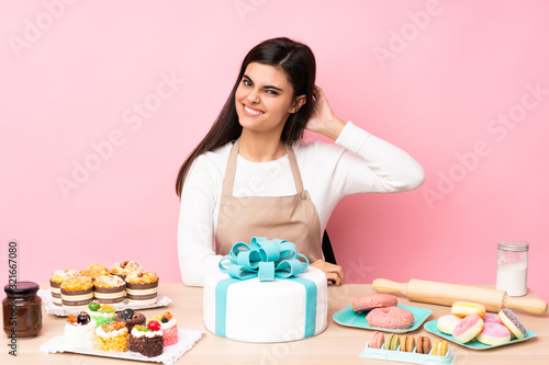 Pastry chef with a big cake in a table over isolated pink background having doubts
