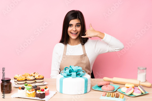 Pastry chef with a big cake in a table over isolated pink background making phone gesture. Call me back sign
