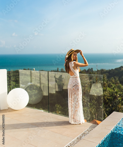 A beautiful girl in a dress is resting in a luxury villa by the pool with wine and in a hat taking cover from the sun. View of palm trees and the sea. Thailand, Koh Samui