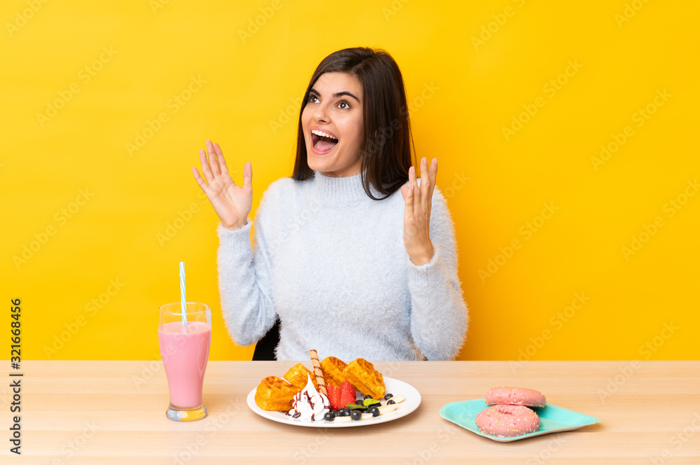 Young woman eating waffles and milkshake in a table over isolated yellow background with surprise facial expression Young woman eating waffles and milkshake in a table over isolated yellow background with surprise facial expression