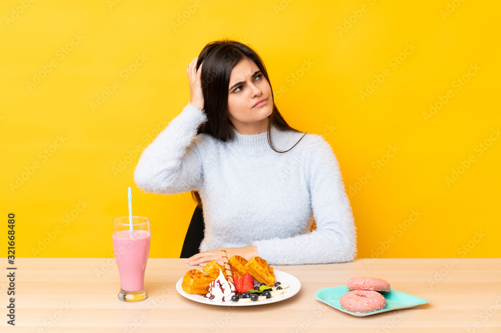 Young woman eating waffles and milkshake in a table over isolated yellow background having doubts and with confuse face expression
