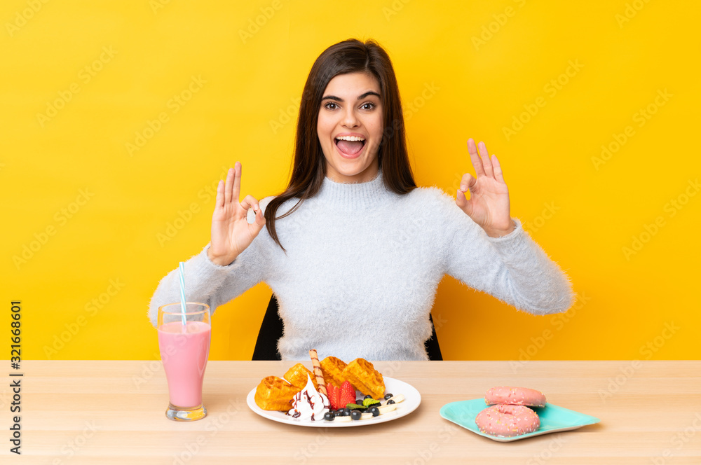 Young woman eating waffles and milkshake in a table over isolated yellow background showing an ok sign with fingers Young woman eating waffles and milkshake in a table over isolated yellow background showing an ok sign with fingers