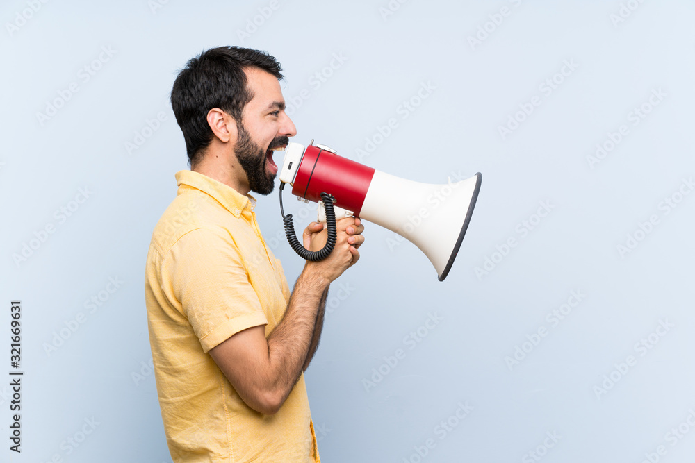Young man with beard over isolated blue background shouting through a megaphone Young man with beard over isolated blue background shouting through a megaphone