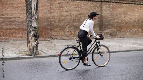 A girl rides a bicycle along the road along the buildings. A girl in a black hat and suspenders