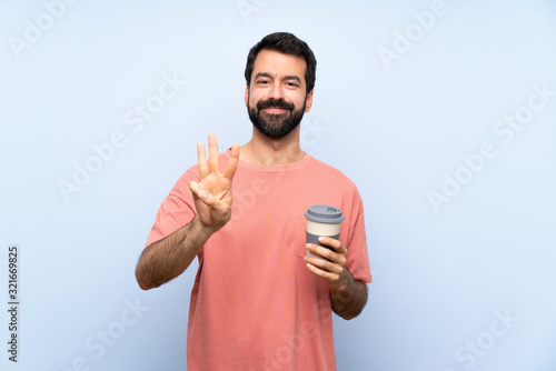 Young man with beard holding a take away coffee over isolated blue background happy and counting three with fingers