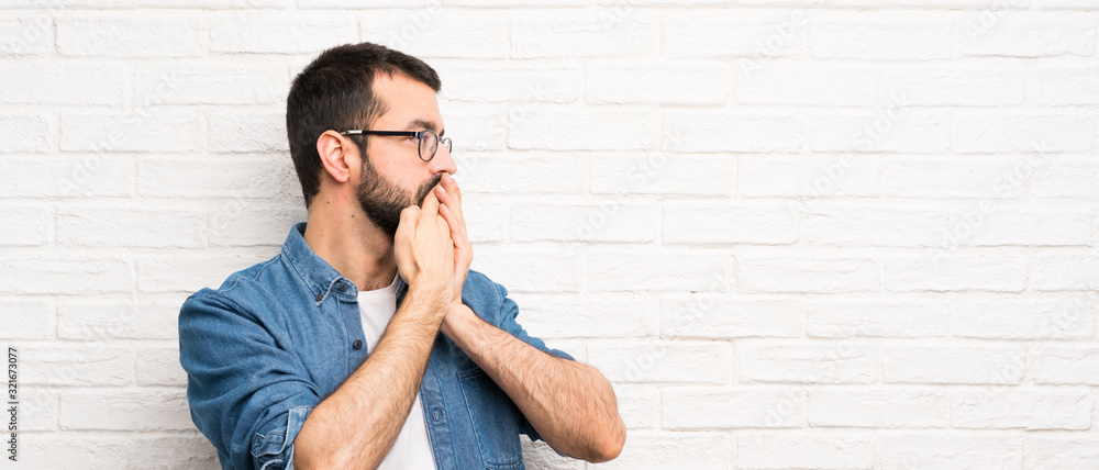 Handsome man with beard over white brick wall covering mouth and looking to the side Handsome man with beard over white brick wall covering mouth and looking to the side
