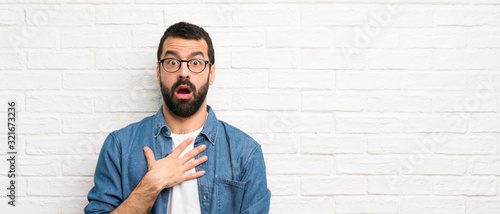 Handsome man with beard over white brick wall surprised and shocked while looking right