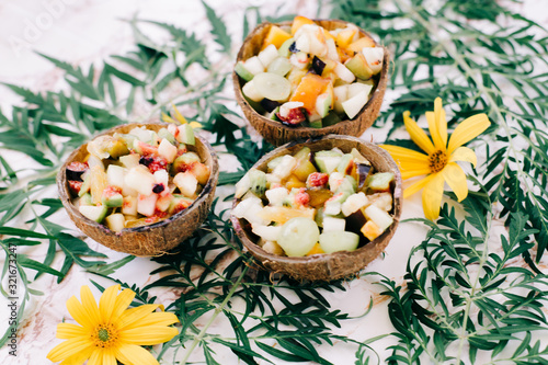 healthy fruit salad in a bowl of coconut shells on the table with tropical leaves. vegetarian food