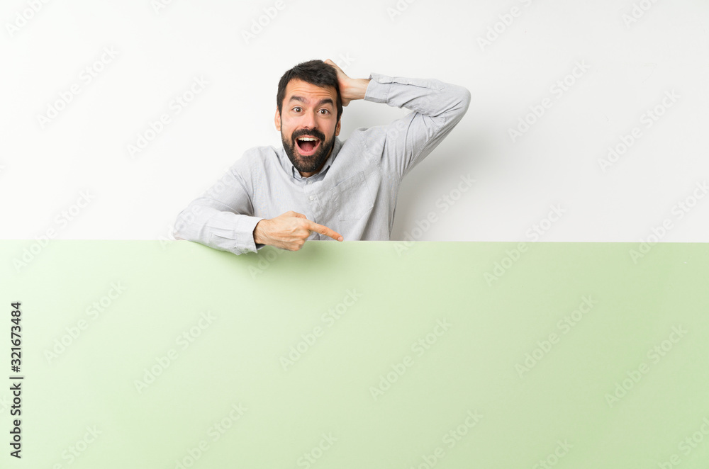 Young handsome man with beard holding a big green empty placard surprised and pointing finger to the side Young handsome man with beard holding a big green empty placard surprised and pointing finger to the side