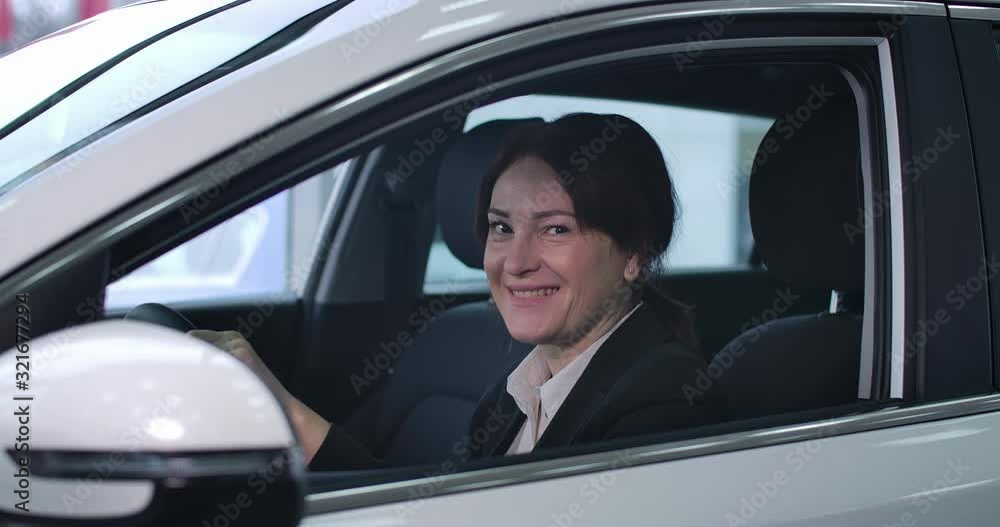 Side view of adult Caucasian woman sitting on driver's seat, looking at camera and smiling. Portrait of confident female owner of new car. Automobile industry, dealership. Cinema 4k ProRes HQ. Side view of adult Caucasian woman sitting on driver's seat, looking at camera and smiling. Portrait of confident female owner of new car. Automobile industry, dealership. Cinema 4k ProRes HQ.