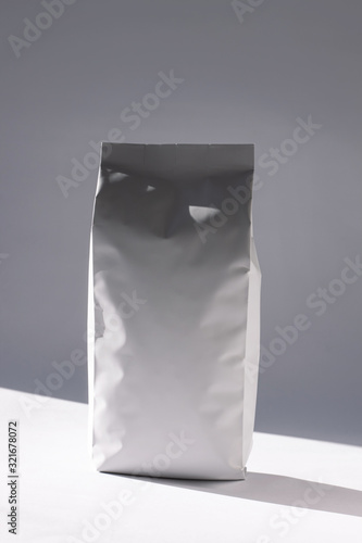 Blank white foil bag on monochrome background in minimal style with natural light shadow. Packaging for foods or drinks with valve and seal template mockup. Metallic coffee tea retail package design.