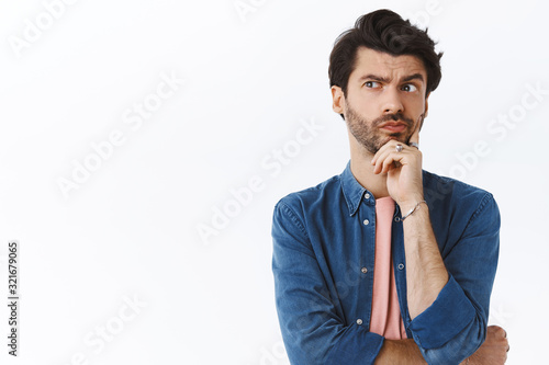 Thoughtful, serious-looking handsome bearded guy, look away pouting pensive, touching chin, thinking, making choice or imaging plan, standing white background pondering how solve situation photo