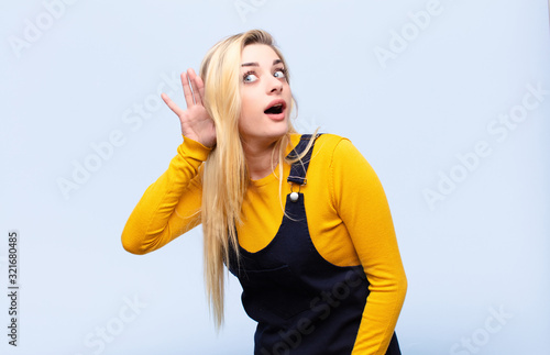 young pretty blonde woman smiling, looking curiously to the side, trying to listen to gossip or overhearing a secret against flat wall photo